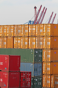 Im Vordergrund bunte Container gestapelt im Hafen, im Hintergrund Kräne