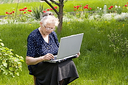 Alte Frau sitzt mit einem Laptop im grünen Garten und tippt
