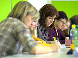 Eine kleine Personengruppe sitzt an einem Tisch, alle schreiben etwas auf Papier