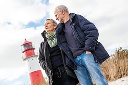 Älteres Paar steht vor einem Leuchtturm am Strand und schaut in die Ferne